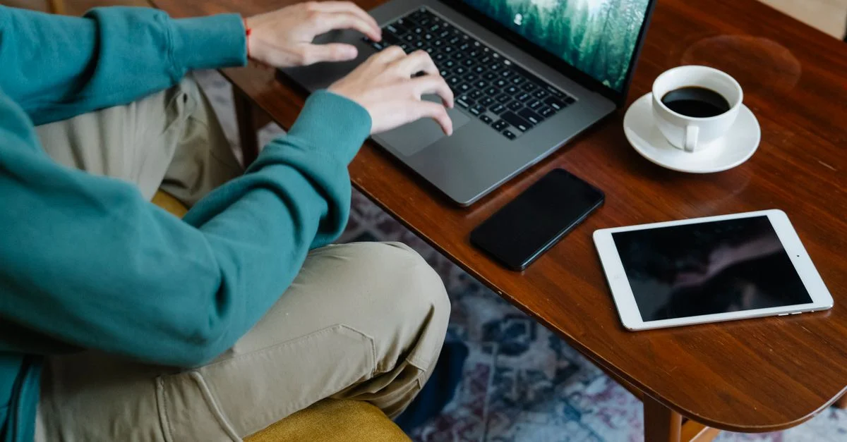 Personne travaillant à distance avec un ordinateur portable, un café, un smartphone et une tablette sur une table en bois.