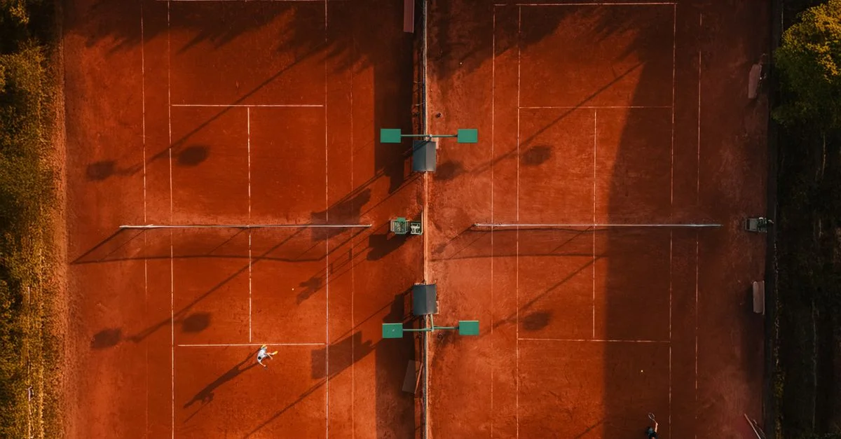 Prise de vue aérienne des courts de tennis en terre battue avec des joueurs pendant l'heure dorée, projetant de longues ombres.